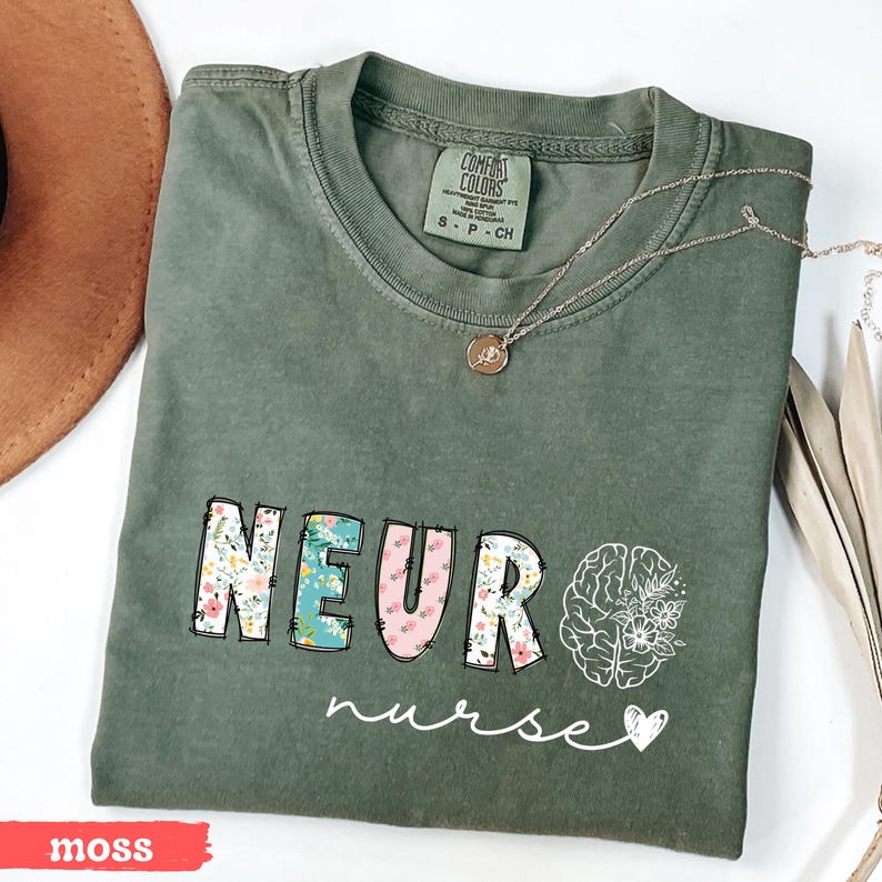 Puede incluir: Una camiseta verde musgo con las palabras "NEURO nurse" en un dise&ntilde;o floral. La palabra "NEURO" tiene un patr&oacute;n floral en las letras. Un collar plateado con un colgante est&aacute; en la camisa.