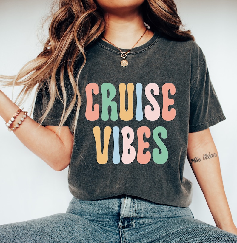 Puede incluir: Una camiseta gris oscuro con el texto "CRUISE VIBES" en letras de estilo retro de colores. Las letras est&aacute;n delineadas en blanco y rellenas de diferentes colores, incluyendo rosa, amarillo, azul y verde.