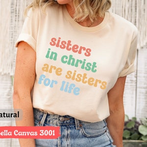 May include: A beige t-shirt with the text "sisters in christ are sisters for life" printed in rainbow colors. The shirt is made by Bella Canvas and is a 3001 style.
