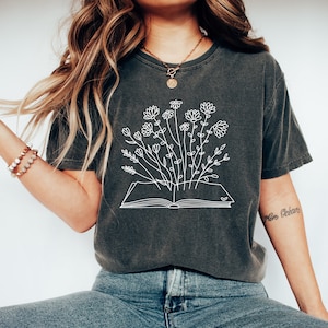 K&ouml;nnte beinhalten: Ein dunkelgraues T-Shirt mit einem wei&szlig;en Umriss eines Buches, aus dem Blumen wachsen. Der Text "More Oceans" befindet sich auf dem rechten &Auml;rmel.
