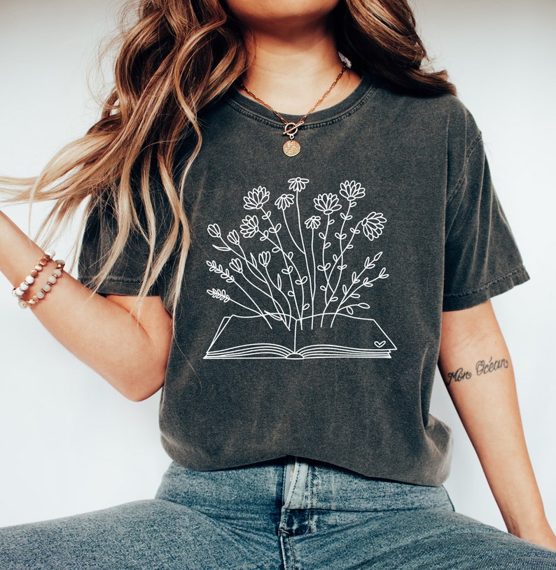 K&ouml;nnte beinhalten: Ein dunkelgraues T-Shirt mit einem wei&szlig;en Umriss eines Buches, aus dem Blumen wachsen. Der Text "More Oceans" befindet sich auf dem rechten &Auml;rmel.