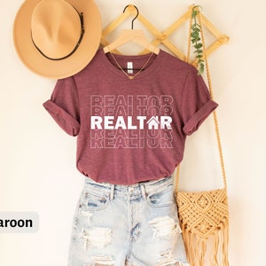 Może przedstawiać: Bordowy t-shirt z napisem "REALTOR" powt&oacute;rzonym w układzie piętrowym z ikoną domu w środku. Koszulka jest zawieszona na drewnianym stojaku z słomkowym kapeluszem i plecionym workiem.