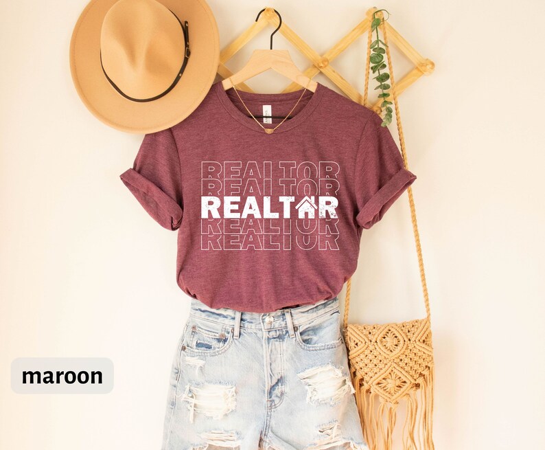 Może przedstawiać: Bordowy t-shirt z napisem "REALTOR" powt&oacute;rzonym w układzie piętrowym z ikoną domu w środku. Koszulka jest zawieszona na drewnianym stojaku z słomkowym kapeluszem i plecionym workiem.