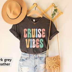 Puede incluir: Camiseta de color gris oscuro jaspeado con el texto "CRUISE VIBES" en letras retro de colores. La camiseta est&aacute; colgada en una percha de madera con un bolso de paja y un sombrero.