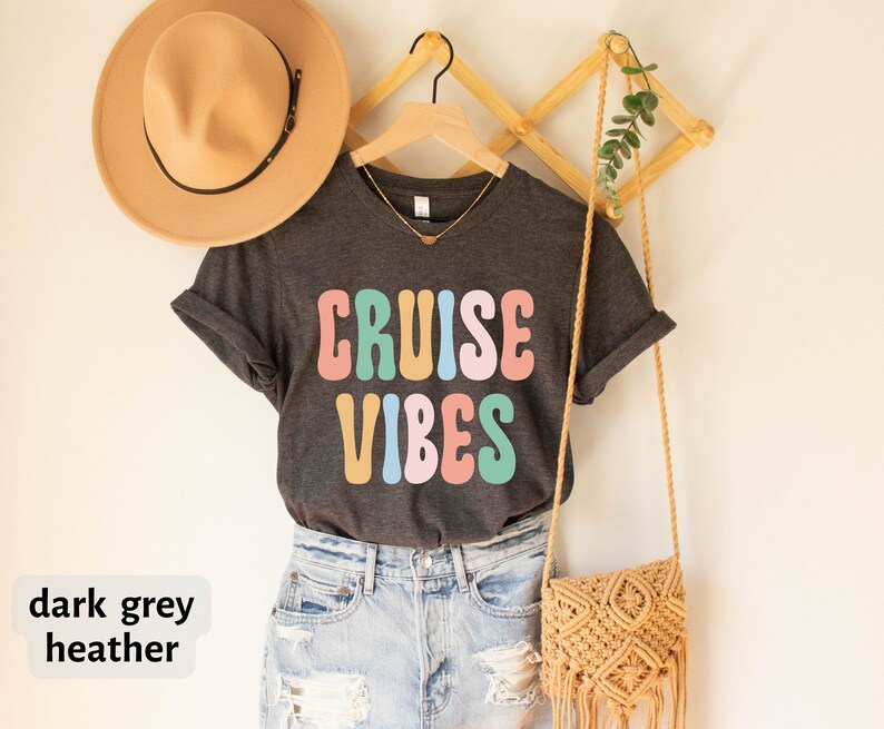 Puede incluir: Camiseta de color gris oscuro jaspeado con el texto "CRUISE VIBES" en letras retro de colores. La camiseta est&aacute; colgada en una percha de madera con un bolso de paja y un sombrero.