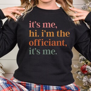 May include: Black sweatshirt with a colourful text graphic that reads "it's me, hi. i'm the officiant, it's me."