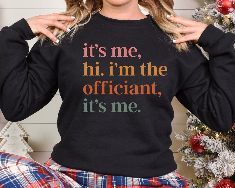 May include: Black sweatshirt with a colourful text graphic that reads "it's me, hi. i'm the officiant, it's me."