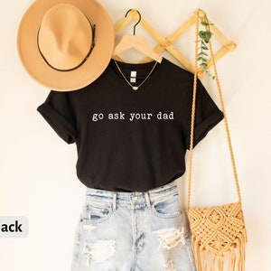 May include: Black t-shirt with the text "go ask your dad" in white lettering. The shirt is hanging on a wooden hanger and is styled with a straw hat and a crocheted bag.