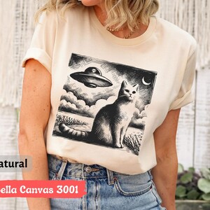 May include: A cream-colored t-shirt with a black and white illustration of a cat and a UFO. The text "Bella Canvas 3001" is printed on the bottom of the shirt.