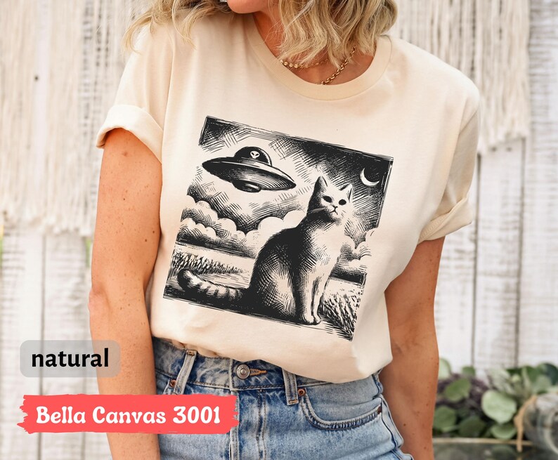 May include: A cream-colored t-shirt with a black and white illustration of a cat and a UFO. The text "Bella Canvas 3001" is printed on the bottom of the shirt.