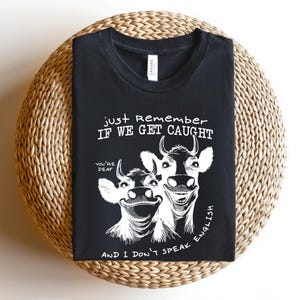 Może przedstawiać: Czarny t-shirt z zabawną grafiką dw&oacute;ch uśmiechniętych kr&oacute;w. Tekst brzmi: "Just Remember If We Get Caught, You're Deaf, And I Don't Speak English." Koszulka jest złożona na okrągłej, plecionej powierzchni.