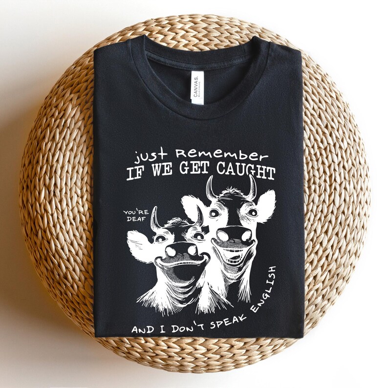Może przedstawiać: Czarny t-shirt z zabawną grafiką dw&oacute;ch uśmiechniętych kr&oacute;w. Tekst brzmi: "Just Remember If We Get Caught, You're Deaf, And I Don't Speak English." Koszulka jest złożona na okrągłej, plecionej powierzchni.
