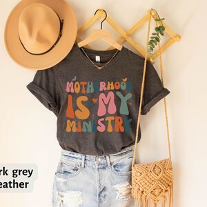May include: Dark grey heather t-shirt with the text "Motherhood is my ministry" in colorful, retro-style lettering.
