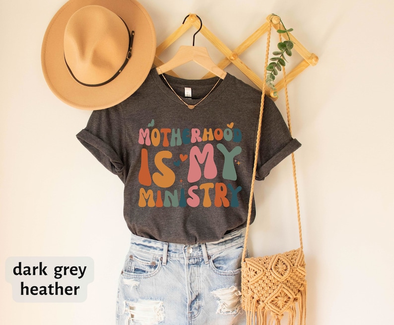 May include: Dark grey heather t-shirt with the text "Motherhood is my ministry" in colorful, retro-style lettering.