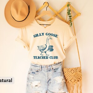 May include: A natural colored t-shirt with a blue graphic of a goose carrying books. The text "Silly Goose Teacher Club" is printed on the shirt.