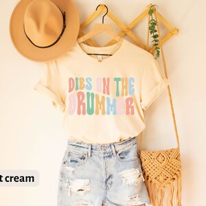 Puede incluir: Una camiseta de color crema suave con el texto "DIBS ON THE DRUMMER" en letras de colores de estilo retro. La camiseta est&aacute; colgada en una percha de madera con un bolso de paja y un sombrero.