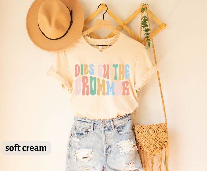 Puede incluir: Una camiseta de color crema suave con el texto "DIBS ON THE DRUMMER" en letras de colores de estilo retro. La camiseta est&aacute; colgada en una percha de madera con un bolso de paja y un sombrero.