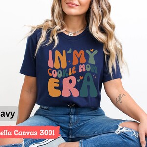 May include: A navy blue t-shirt with the text "In My Cookie Mom Era" in colorful, retro-style lettering. The shirt is made by Bella Canvas and is a 3001 style.