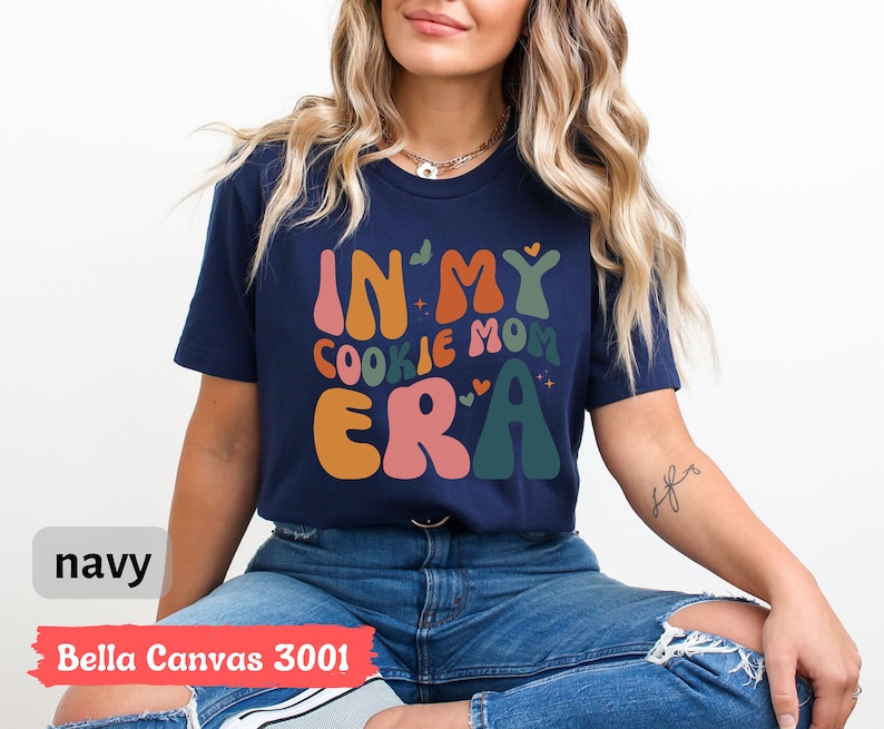 May include: A navy blue t-shirt with the text "In My Cookie Mom Era" in colorful, retro-style lettering. The shirt is made by Bella Canvas and is a 3001 style.