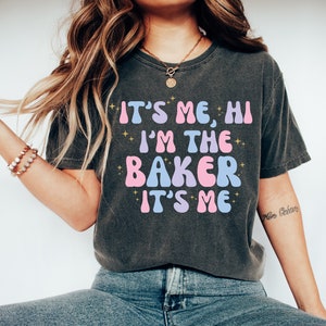 Peut inclure: Un t-shirt gris fonc&eacute; avec le texte "It's Me, Hi I'm The Baker It's Me" en lettres roses, bleues et dor&eacute;es. Le texte est dans un style de police r&eacute;tro.