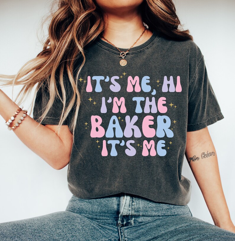 Peut inclure: Un t-shirt gris fonc&eacute; avec le texte "It's Me, Hi I'm The Baker It's Me" en lettres roses, bleues et dor&eacute;es. Le texte est dans un style de police r&eacute;tro.