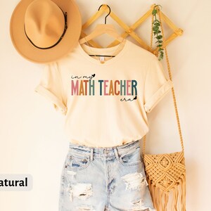 May include: A beige t-shirt with the text "in my heart math teacher era" in rainbow colours. The shirt is hanging on a wooden hanger with a straw bag and a hat.