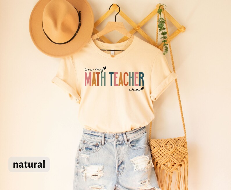 May include: A beige t-shirt with the text "in my heart math teacher era" in rainbow colours. The shirt is hanging on a wooden hanger with a straw bag and a hat.