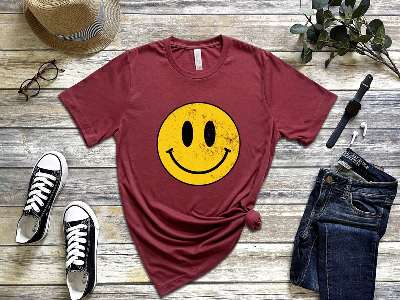 May include: A maroon t-shirt featuring a yellow smiley face graphic. The smiley face has a distressed, vintage look.