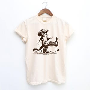 May include: Cream-colored t-shirt featuring a brown illustration of a cat wearing a cowboy hat and boots, walking across a desert scene with cacti. The shirt is hanging on a wooden hanger.