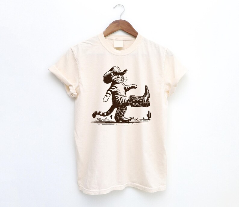 May include: Cream-colored t-shirt featuring a brown illustration of a cat wearing a cowboy hat and boots, walking across a desert scene with cacti. The shirt is hanging on a wooden hanger.