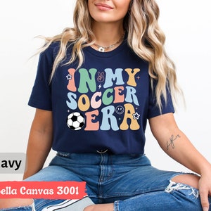 Może przedstawiać: Koszulka w kolorze granatowym z napisem "In My Soccer Era" w kolorowych, retro literach. Projekt zawiera piłkę nożną, znak pokoju, błyskawicę i uśmiechniętą buźkę.