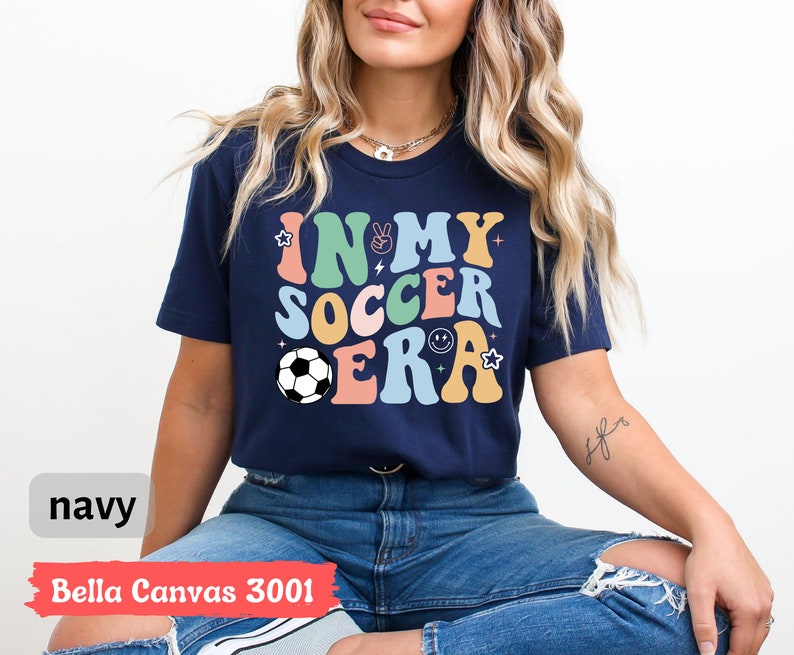Może przedstawiać: Koszulka w kolorze granatowym z napisem "In My Soccer Era" w kolorowych, retro literach. Projekt zawiera piłkę nożną, znak pokoju, błyskawicę i uśmiechniętą buźkę.