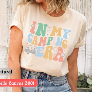 May include: A beige t-shirt with the text "In My Camping Era" in colorful retro font. The text is surrounded by stars and a small camper van graphic.