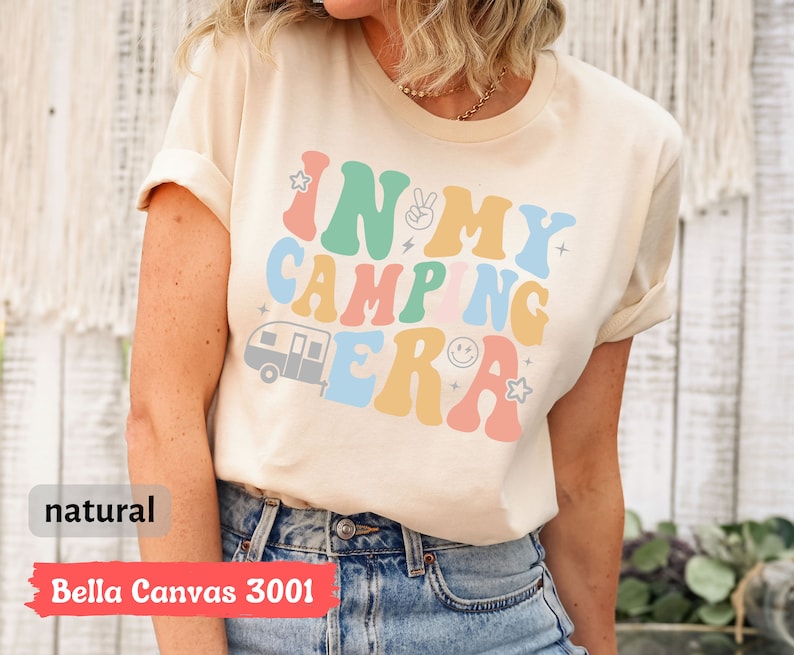 May include: A beige t-shirt with the text "In My Camping Era" in colorful retro font. The text is surrounded by stars and a small camper van graphic.