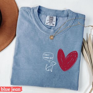 May include: A blue jean Comfort Colors t-shirt with a white cartoon dog and a red heart graphic. The dog has a speech bubble that says "Can I come in?" A gold necklace is draped across the shirt.