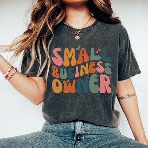 May include: A dark gray t-shirt with the text "SMALL BUSINESS OWNER" in colorful, retro-style lettering. The letters are outlined in black and have small hearts and stars around them.