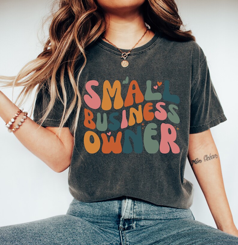 May include: A dark gray t-shirt with the text "SMALL BUSINESS OWNER" in colorful, retro-style lettering. The letters are outlined in black and have small hearts and stars around them.