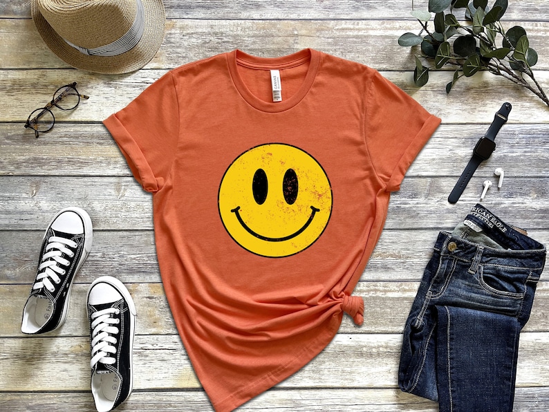 May include: A burnt orange t-shirt featuring a yellow smiley face graphic. The smiley face has a distressed look with a black outline.