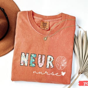 Puede incluir: Una camiseta de color coral con las palabras "NEURO nurse" en un dise&ntilde;o floral. Un collar dorado y un sombrero marr&oacute;n est&aacute;n al lado de la camiseta. Un gr&aacute;fico de cerebro blanco est&aacute; en el lado derecho de la camiseta.