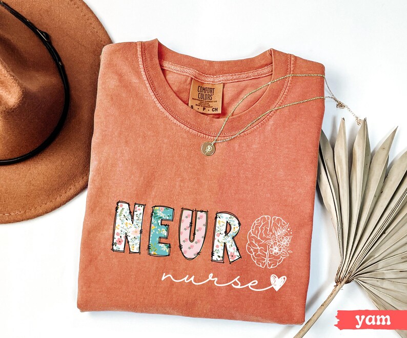 Puede incluir: Una camiseta de color coral con las palabras "NEURO nurse" en un dise&ntilde;o floral. Un collar dorado y un sombrero marr&oacute;n est&aacute;n al lado de la camiseta. Un gr&aacute;fico de cerebro blanco est&aacute; en el lado derecho de la camiseta.