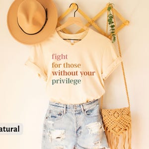 Op de afbeelding: Een beige T-shirt met de tekst "fight for those without your privilege" gedrukt in verschillende tinten roze, oranje en groen. Het shirt hangt aan een houten rek met een rieten tas en een hoed.
