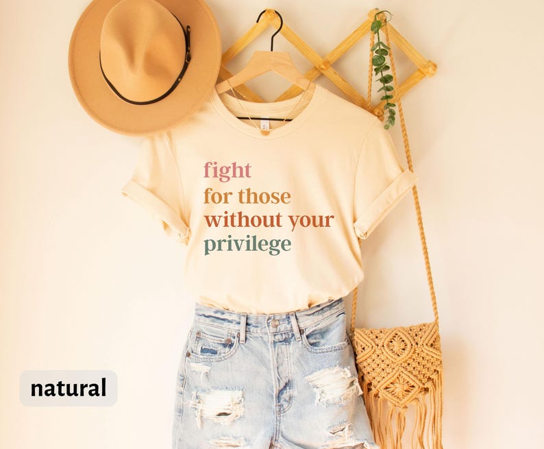 Op de afbeelding: Een beige T-shirt met de tekst "fight for those without your privilege" gedrukt in verschillende tinten roze, oranje en groen. Het shirt hangt aan een houten rek met een rieten tas en een hoed.