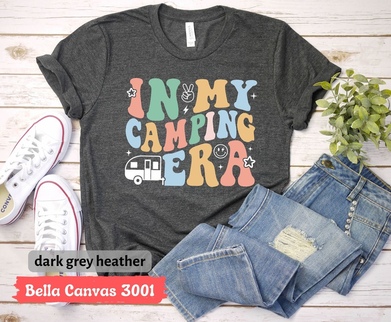 May include: A dark grey heather Bella Canvas 3001 t-shirt with the text "In My Camping Era" in colorful retro letters. A small camper van graphic is below the text.