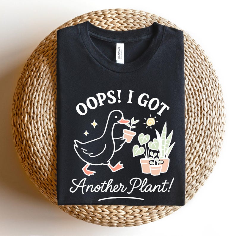 May include: Black t-shirt with the text "OOPS! I GOT Another Plant!" and a graphic of a duck carrying a potted plant. Additional graphics include a sun, stars, and potted plants. The shirt is folded on a woven, circular surface.