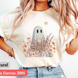 Pu&ograve; includere: Una maglietta color crema con un fantasioso disegno di fantasma in mezzo a un campo di fiori selvatici nei toni dell'arancione, del rosa e del viola. Una luna piena &egrave; sullo sfondo. La maglietta &egrave; etichettata "natural" e "Bella Canvas 3001".