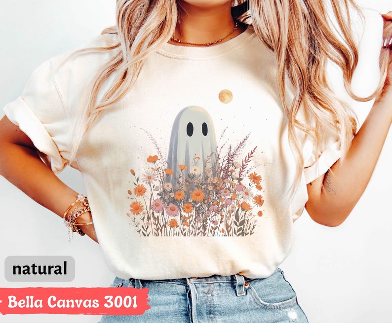 Pu&ograve; includere: Una maglietta color crema con un fantasioso disegno di fantasma in mezzo a un campo di fiori selvatici nei toni dell'arancione, del rosa e del viola. Una luna piena &egrave; sullo sfondo. La maglietta &egrave; etichettata "natural" e "Bella Canvas 3001".