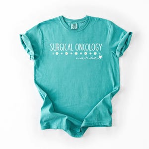 May include: Teal blue t-shirt with the text "SURGICAL ONCOLOGY nurse" in white lettering with small white flowers and a heart.
