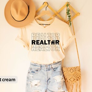 Może przedstawiać: Jasno kremowy t-shirt z tekstem "REALTOR" powt&oacute;rzonym cztery razy w kolorze czarnym, z ikoną domu w drugim "REALTOR".