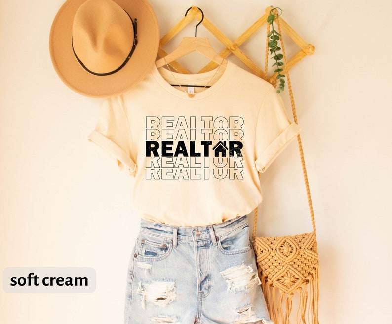 Może przedstawiać: Jasno kremowy t-shirt z tekstem "REALTOR" powt&oacute;rzonym cztery razy w kolorze czarnym, z ikoną domu w drugim "REALTOR".