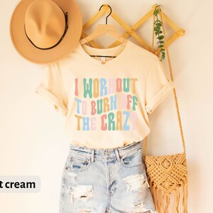 May include: A soft cream colored t-shirt with the text "I workout to burn off the crazy" in colorful, retro-style lettering.